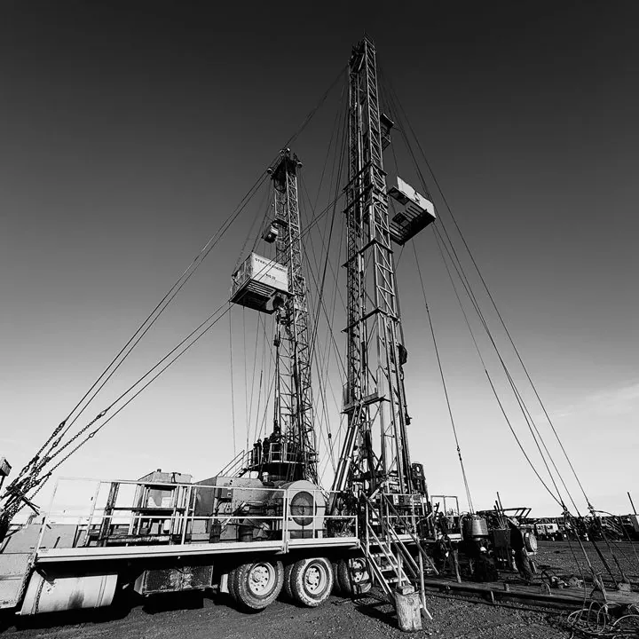 Black and white photo of two well service rigs on location, derricks up and guyed out, with the rig carrier trucks in front