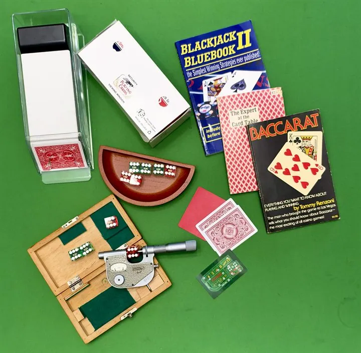 Casino gear on green felt: a card shoe, dice in a bowl and in a wooden case with a dice micrometer, playing cards, a craps strategy card, and books on blackjack and baccarat